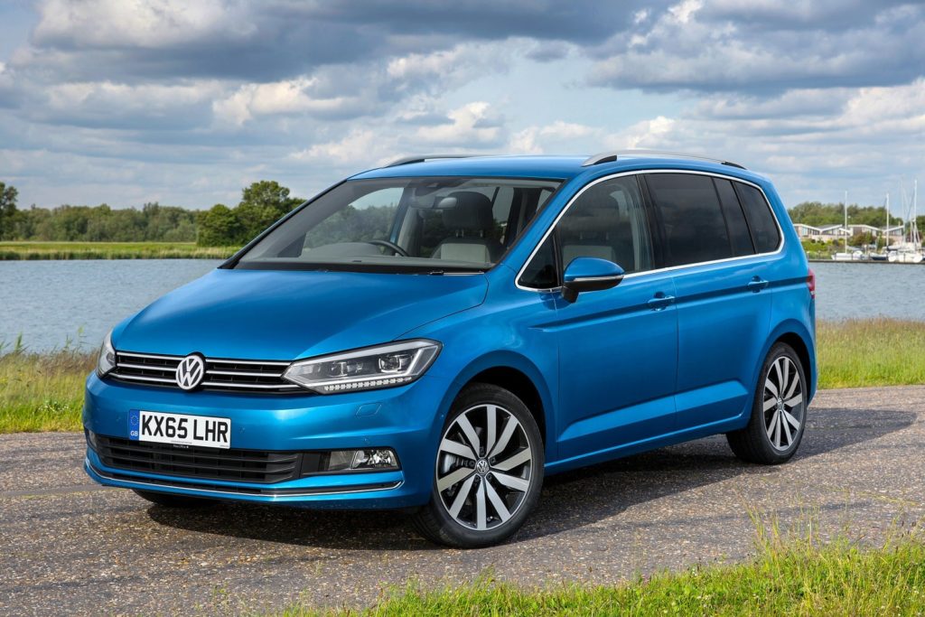 a metallic blue volkswagen touran parked on tarmac with green grass and the sea in the background