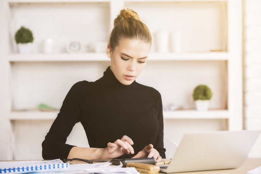 lady in black turtleneck with blonde hair up in a ponytail works something out on her phone and laptop