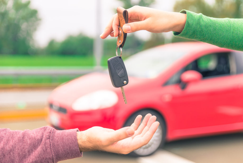 person with a green top on handing the keys to a car to someone in a purple top with a red car in the background