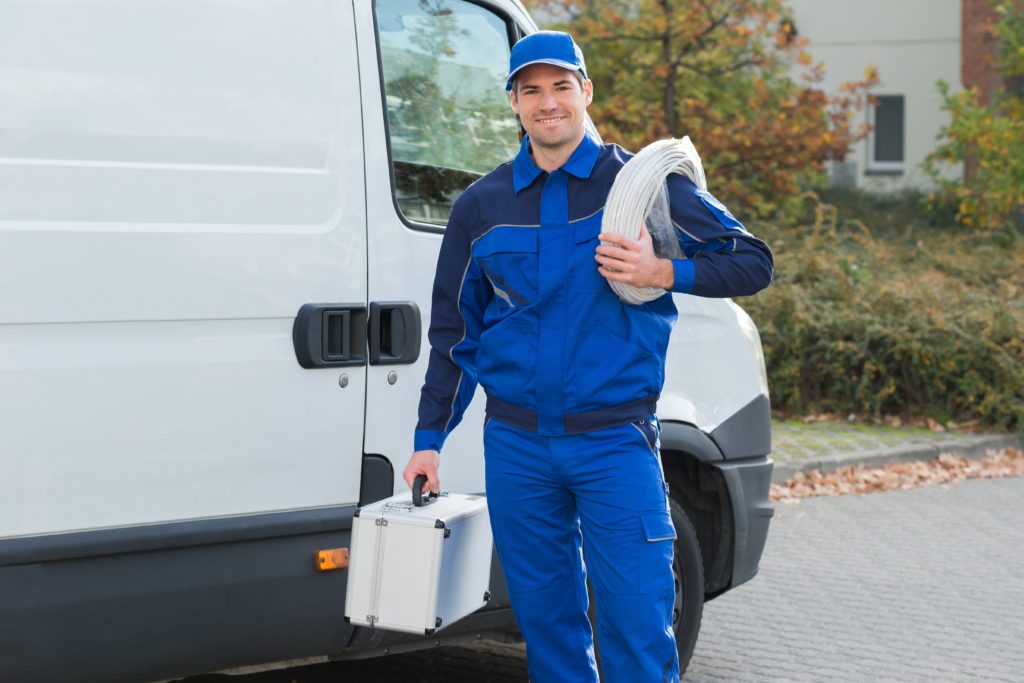 An electrician in a blue jump suit carrying roll of white cable standing next to his white van