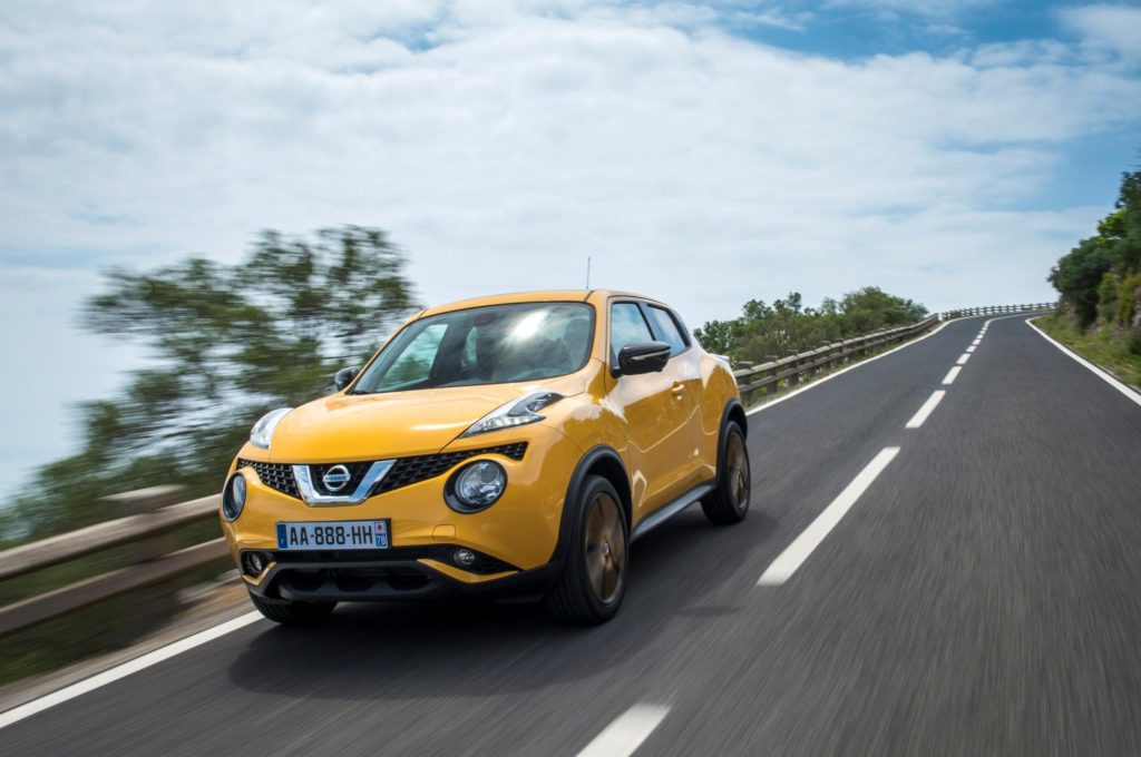 bright yellow nissan juke hatchback driving on tarmac road with green trees in the background and blue skies in the background