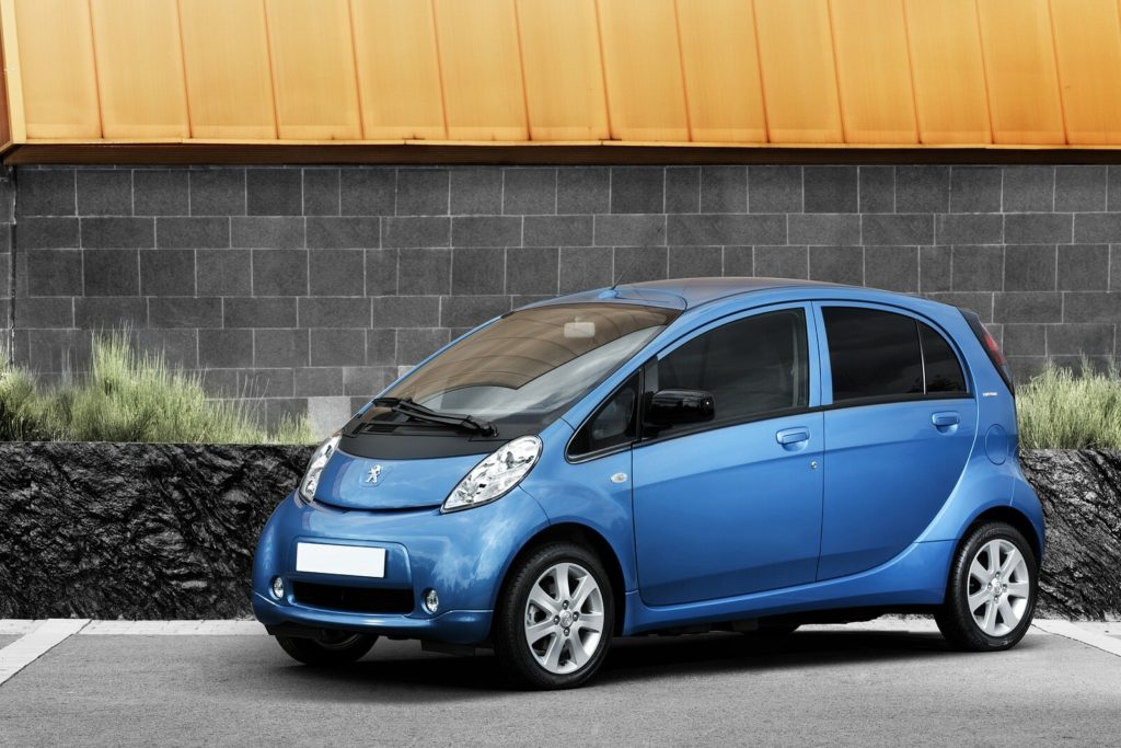 a metallic blue peugeot ion hatchback parked on tarmac infront of bright building