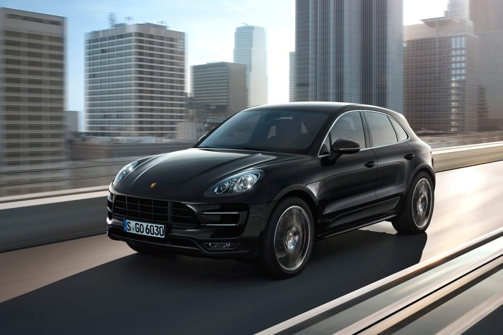 a dark porsche macan estate driving on tarmac road with a big city in the background