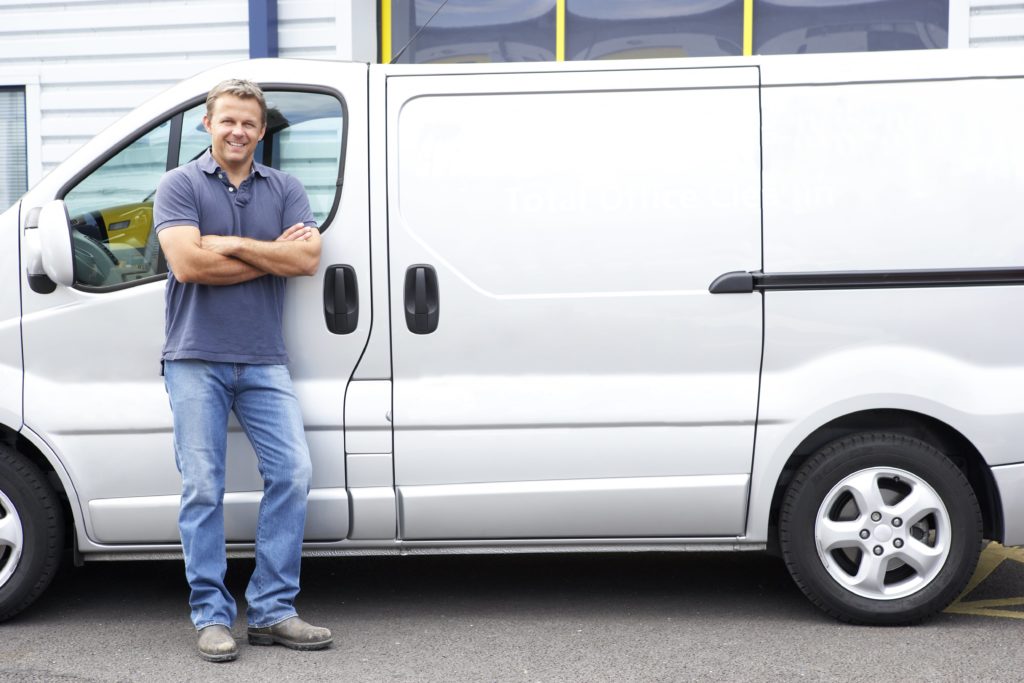 man in a blue polo shirt and blue jeans standing in front of a silver mid sized van smiling