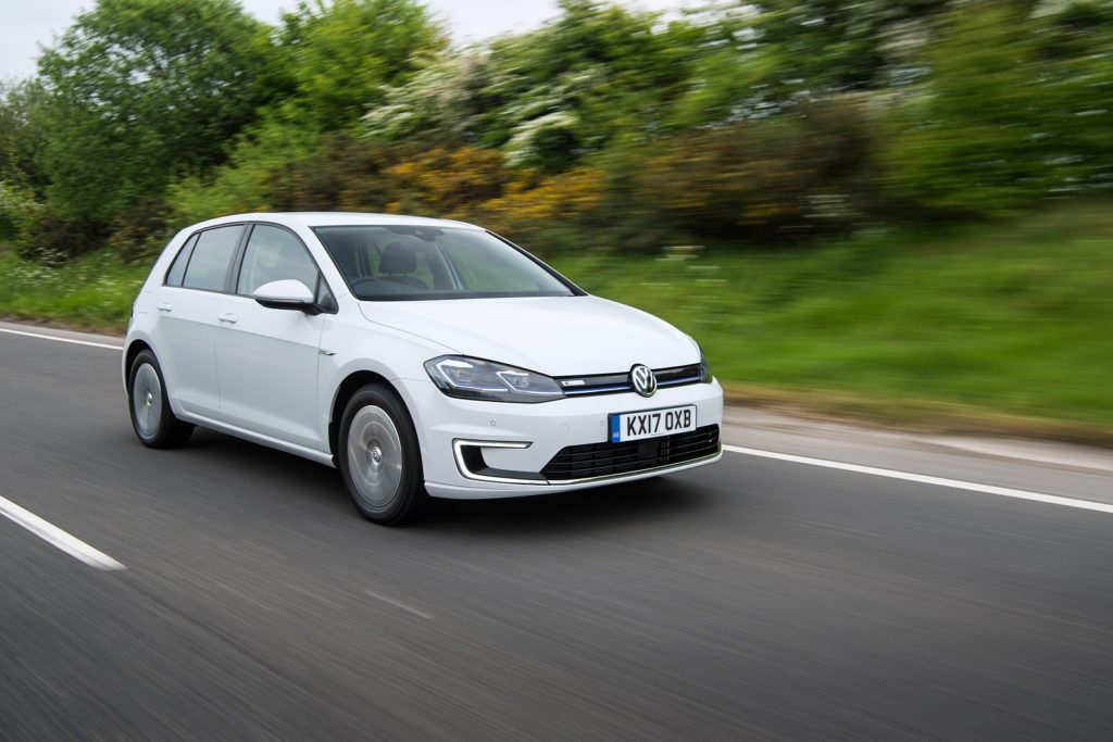 a metallic white volkswagen e golf driving on tarmac road with green bushes in the backrground